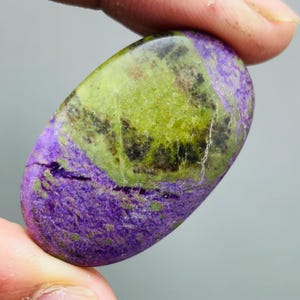 Peut inclure: Une pierre précieuse ovale à surface polie. La pierre présente une combinaison de couleurs saisissante de violet et de vert, avec un mélange de textures et de motifs. La pierre est tenue entre deux doigts.