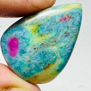 May include: A polished gemstone with a mix of colours. The stone features a vibrant magenta inclusion set within a matrix of blue, green, and yellow hues. The stone is held between two fingers.