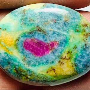 May include: A polished gemstone with a vibrant mix of colours. The stone features a central, vivid pink inclusion surrounded by a blend of teal, yellow, and green hues. The stone's surface is smooth and rounded.