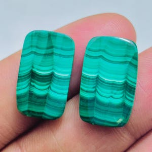 May include: Two polished malachite gemstones. Each stone is rectangular with rounded corners and displays a vibrant green color with dark green, wave-like patterns. The gemstones are held in a hand.