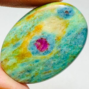 Könnte beinhalten: Ovaler Edelstein mit einer lebendigen Farbmischung. Der Stein hat eine Basis aus Türkis und Gelb, mit auffälligen magenta- und blaufarbenen Einschlüssen. Die polierte Oberfläche hebt die natürlichen Muster und Texturen des Steins hervor.