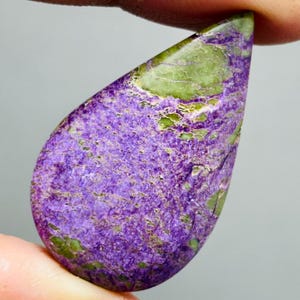 Peut inclure: Une pierre précieuse en forme de goutte d'eau avec une surface texturée. La pierre présente un mélange de teintes violettes et vertes, le violet dominant la partie inférieure et le vert le haut. La pierre est tenue entre deux doigts.