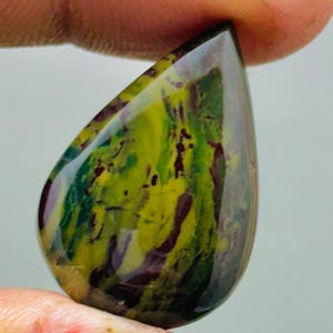 Könnte beinhalten: Ein tropfenförmiger, polierter Edelstein. Der Stein zeigt eine Mischung aus Farben, darunter Gelb, Grün und tiefrote-violette Farbtöne. Der Edelstein wird zwischen zwei Fingern gehalten, was seine Größe und Form hervorhebt.