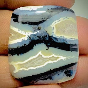 Op de afbeelding: Een vierkante edelsteen met een gelaagd patroon van zwart, wit, grijs en lichtgeel. De steen heeft een gepolijst oppervlak en een natuurlijk uiterlijk. De lagen creëren een uniek, abstract ontwerp.