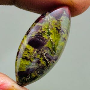 Könnte beinhalten: Ein oval geformter Edelstein mit polierter Oberfläche. Der Stein weist eine Mischung aus Farben auf, darunter tiefes Lila, leuchtendes Grün und Hauch von Rot. Der Edelstein wird zwischen zwei Fingern gehalten.