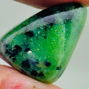 Könnte beinhalten: Ein polierter, dreieckiger Edelstein mit leuchtend grüner Farbe, gesprenkelt mit schwarzen Einschlüssen. Der Stein hat eine glatte, glänzende Oberfläche und wird zwischen zwei Fingern gehalten. Die Form des Edelsteins erinnert an eine kleine, abgerundete Pyramide.