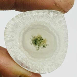 May include: A teardrop-shaped gemstone with translucent white and grey bands. The centre has a cluster of green, moss-like inclusions. The stone is held between fingers, highlighting its natural beauty and unique patterns.
