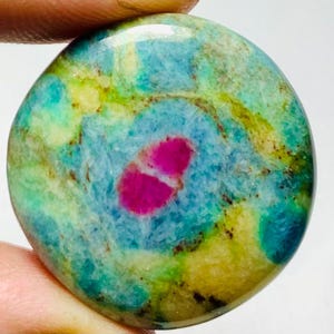 May include: A circular gemstone with a polished surface. The stone displays a central area of vivid pink, encircled by swirling patterns of blue, green, and yellow. The gemstone is held between fingers.
