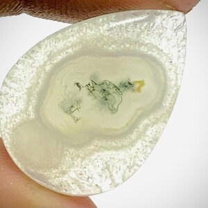 Puede incluir: Una piedra preciosa en forma de lágrima con una base translúcida blanquecina. La piedra presenta anillos concéntricos de diferentes tonos de blanco y una zona central con inclusiones verdes y amarillas. La piedra se sujeta entre dos dedos.