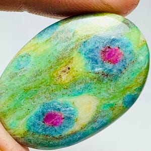 May include: An oval gemstone with a striking colour scheme. The stone displays a base of green and yellow tones, with patches of blue and vibrant pink centres. The polished surface reflects light, emphasising the natural patterns.