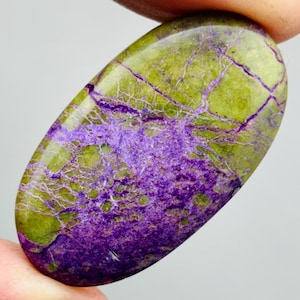 Pode incluir: Gema oval com superfície polida. A pedra apresenta uma mistura de cores, incluindo verde oliva e roxo profundo, com uma rede de finas linhas contrastantes. A gema é segurada entre os dedos.