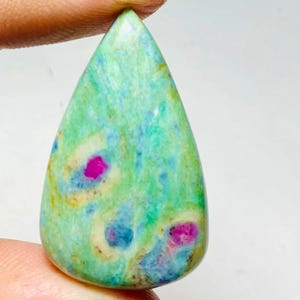 May include: A teardrop-shaped gemstone with a vibrant mix of colours. The stone features a base of light green with swirls of blue and yellow. Bright pink spots are scattered throughout the stone, adding a pop of colour. The gemstone is held between two fingers.