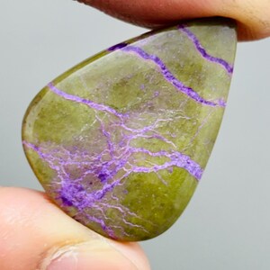 Könnte beinhalten: Ein polierter Edelstein in Tropfenform. Der Stein hat eine olivgrüne Grundfarbe mit auffälligen violetten Adern. Der Stein wird zwischen zwei Fingern gehalten, was seine glatte, glänzende Oberfläche zeigt. Ein Schmuckstück oder Dekorationsartikel.