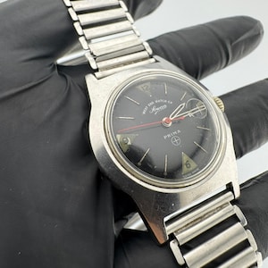 May include: A vintage silver-toned wristwatch with a black face. The watch features the text "West End Watch Co. Sowar Prima" and "Swiss Made". The watch has a silver band and a round face with silver and red hands.