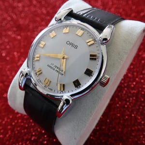 May include: A vintage Oris wristwatch with a silver-colored face, gold-toned hands and markers, and a black leather band. The watch face displays the brand name and text indicating 17 jewels and shock proof. The watch is set on a white surface against a red background.