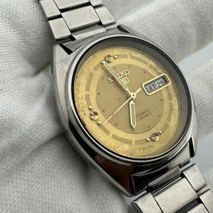 May include: A vintage Seiko automatic wristwatch with a gold-toned face and a silver stainless steel strap. The watch face displays the day and date, with the day reading "SAT 29". The watch has a classic design.