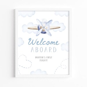 May include: A framed print featuring a watercolor illustration of a biplane with a blue propeller and clouds. The text "Welcome Aboard" and "Martin's First Flight!" are written in blue script.