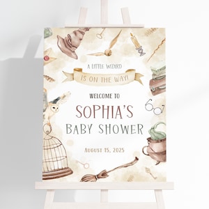 May include: A watercolor baby shower sign on an easel. The sign features a wizard theme with a banner that reads "A Little Wizard Is On The Way!" and "Welcome to Sophia's Baby Shower" with the date August 15, 2025. The design includes a wizard hat, owl, and broom.