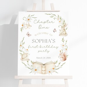 May include: A white sign on an easel with a floral wreath design. The sign reads "Chapter One, welcome to SOPHIA'S first birthday party" with the date March 24, 2026. The wreath includes daisies, butterflies, and woodland creatures.