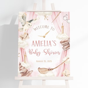 May include: A pink and white baby shower sign on an easel. The sign features a watercolor design with an owl, broom, and other magical elements. The text reads "Welcome to Amelia's Baby Shower" and the date August 15, 2025.