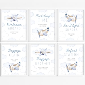 May include: Six framed prints with a watercolor airplane theme. Each print features a light blue airplane, clouds, and text such as "Welcome Aboard" and "Baggage Claim". The prints are in a soft, pastel color palette.