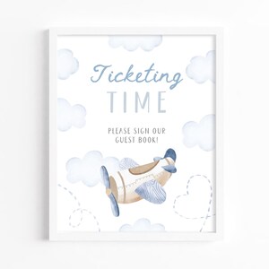 May include: A framed print with a watercolor illustration of a vintage airplane and clouds. The text reads "Ticketing Time, Please Sign Our Guest Book!" in blue and gray lettering. The artwork has a soft, pastel color palette.
