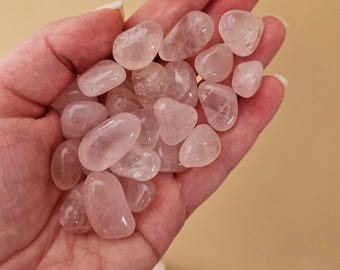 May include: A handful of polished rose quartz stones. The stones are a soft pink color, smooth, and rounded. They vary in size and shape, with a translucent quality. The stones are held in a hand against a neutral background.