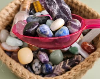 May include: A bright pink plastic scoop filled with a variety of polished and raw gemstones, including amethyst, sodalite, and quartz, held over a woven basket containing a larger collection of assorted colorful tumbled stones.