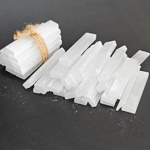 May include: A collection of white selenite crystal sticks. Some are bundled with twine, while others are scattered. The crystals have a translucent appearance and a rectangular shape. The background is a dark grey.