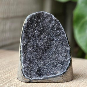 May include: A dark grey geode crystal with a sparkling interior. The crystal has a rough, natural exterior and a cavity filled with tiny, glittering crystals. The geode is displayed upright, showcasing its unique texture and colour variations.