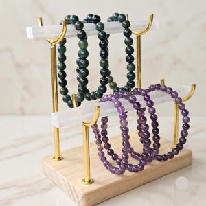 May include: A two-tiered jewellery display stand with a light wood base, gold metal supports, and two horizontal white bars. Three dark green beaded bracelets hang from the top bar, and three purple beaded bracelets hang from the bottom bar.