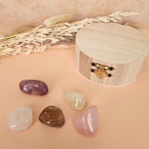 May include: A wooden, round box with a brass clasp sits next to a collection of polished gemstones. The gemstones are various colors, including purple, clear, brown, and pink. Dried plants are in the background.