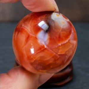 May include: A polished, spherical carnelian gemstone displaying deep orange and reddish-brown hues with translucent white inclusions and natural internal fractures. The smooth, rounded surface reflects light, highlighting the varied colour patterns.