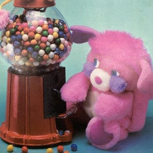 May include: A vintage coloring book titled "The Popples' Magic Gum Ball Machine" featuring a colorful illustration. The cover shows a gumball machine with a plush toy on top and another to the side. The background is a light blue.