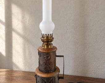May include: A table lamp featuring a tall white glass chimney, a brass burner, and a base constructed from two stacked cylindrical wooden sections wrapped in dark metal plates with embossed fleur-de-lis and geometric patterns.