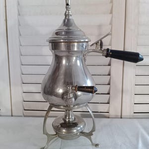 Peut inclure: Une cafetière vintage argentée avec un corps bulbeux, un bec verseur et une poignée en bois noir. La cafetière repose sur un support à trois pieds. Le dessus a une caractéristique en forme de sifflet.