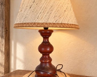 May include: A table lamp with a dark brown turned wood base and a conical, textured beige fabric lampshade featuring a decorative braided trim along the bottom edge. The lamp includes a brown electrical cord with an inline rocker switch.