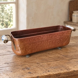 Pode incluir: Uma jardineira retangular de cobre martelado com acabamento metálico. O recipiente possui duas alças laterais com detalhes em cerâmica branca e azul e apoia-se sobre quatro pequenos pés de metal texturizado. O interior é liso e profundo.