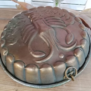 Puede incluir: Molde de pastel antiguo de color bronce con un diseño floral detallado. El molde redondo tiene un borde festoneado y un pequeño anillo para colgar. El metal envejecido tiene una pátina, lo que sugiere su antigüedad.