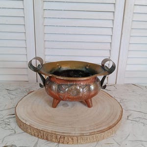Può includere: Una piccola ciotola antica in rame con interno scuro e design floreale in rilievo. La ciotola ha tre piccole gambe e due manici curvi. Si trova su una fetta di legno rotonda, con uno sfondo bianco a persiane.