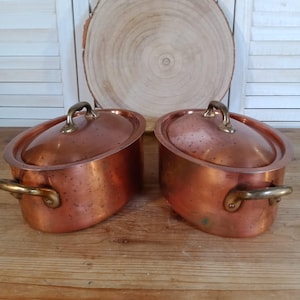 Pode incluir: Duas panelas ovais de cobre com tampas e alças de latão. As panelas são de uma cor marrom-avermelhada quente, com um acabamento brilhante. As tampas têm uma forma curva e são encimadas por uma alça de latão. As panelas são provavelmente para cozinhar.