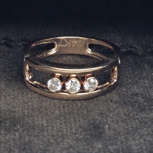 May include: Rose gold ring featuring three clear, round gemstones. The ring has an open design with cut-out details. The ring is displayed on a dark grey, textured surface.