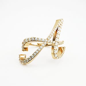Puede incluir: Un anillo de oro con forma de letra A, que presenta un acabado metálico pulido y una serie de pequeñas piedras redondas transparentes incrustadas a lo largo de toda la superficie frontal de la letra, creando un efecto brillante continuo.