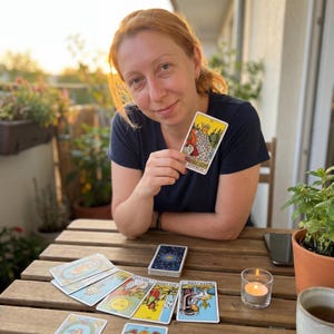 May include: A person in a dark blue t-shirt holding a tarot card labeled The Empress, with several other tarot cards spread out on a wooden table, including cards depicting the sun, a queen, and a world scene, alongside a small lit tea light candle.