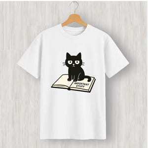 May include: White t-shirt with a black cat illustration sitting on an open book. The book has the words "IMPORTANT STUFF" printed on it. The cat has a grumpy expression. The t-shirt is hanging on a wooden hanger.