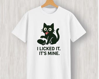 May include: White t-shirt featuring a black cartoon cat with its tongue sticking out, sitting next to a dark object. The text "I LICKED IT. IT'S MINE." is printed below the cat in a bold font. The shirt is hanging on a wooden hanger.