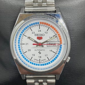 Könnte beinhalten: Eine silberfarbene Seiko Automatik-Armbanduhr mit weißem Zifferblatt, blauen und orangefarbenen Akzenten und einem Edelstahlarmband. Das Zifferblatt zeigt den Tag und das Datum an, mit dem Wort "Automatic".