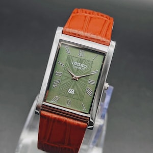 May include: A rectangular Seiko Quartz watch with a silver case and a green face featuring Roman numerals. The watch has a brown leather strap. The watch face has the Seiko Quartz logo and a decorative pattern.