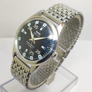 Może przedstawiać: Srebrny zegarek z czarną tarczą i bransoletą ze stali nierdzewnej. Tarcza zegarka posiada białe cyfry i niebieskie akcenty, z napisem "hmt PILOT" i "PARA SHOCK 17 JEWELS". Bransoleta jest w stylu plecionym.