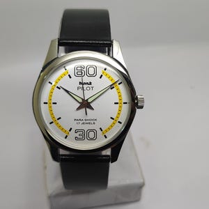 May include: Silver wristwatch with a white face, black leather strap, and a yellow and black bezel. The watch face features the text "hmt PILOT" and "PARA SHOCK 17 JEWELS". The watch has a second hand and a crown.